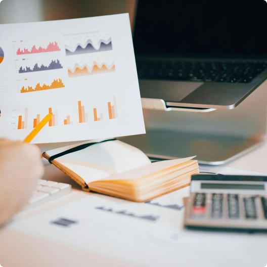 Graphs and charts on paper with orange pencil, notebook, calculator, and laptop on a desk.