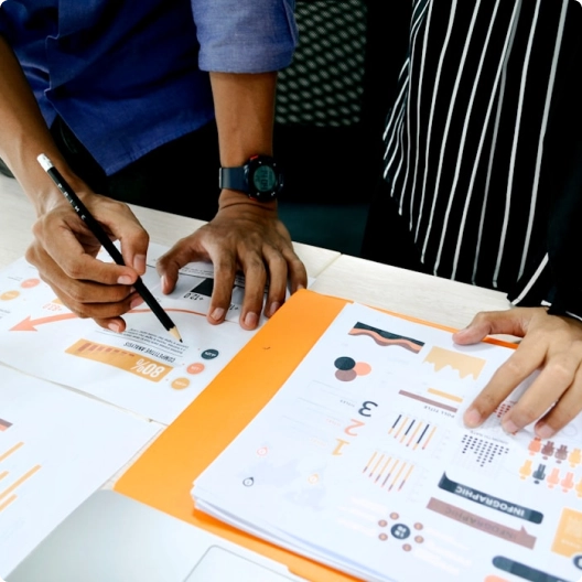 Hands of two people working on colorful printed papers with graphs and notes on a desk.