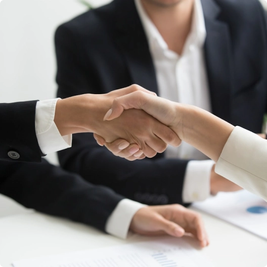 Hands shaking over a table with documents, two people in business attire in the background.
