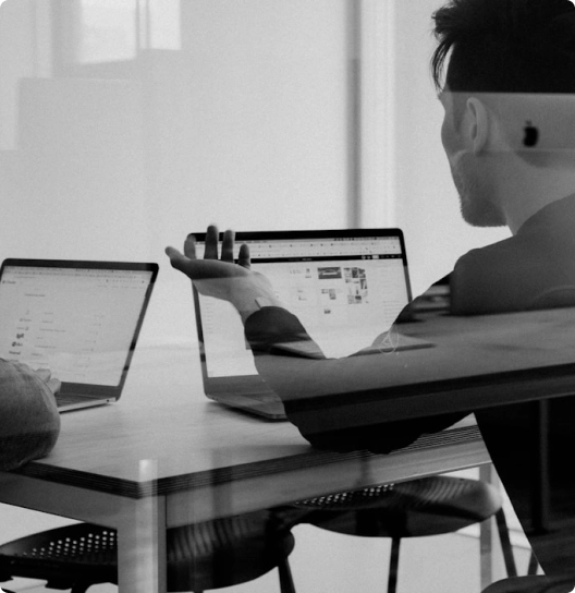 Two people discussing at a table with laptops open in a bright workspace.